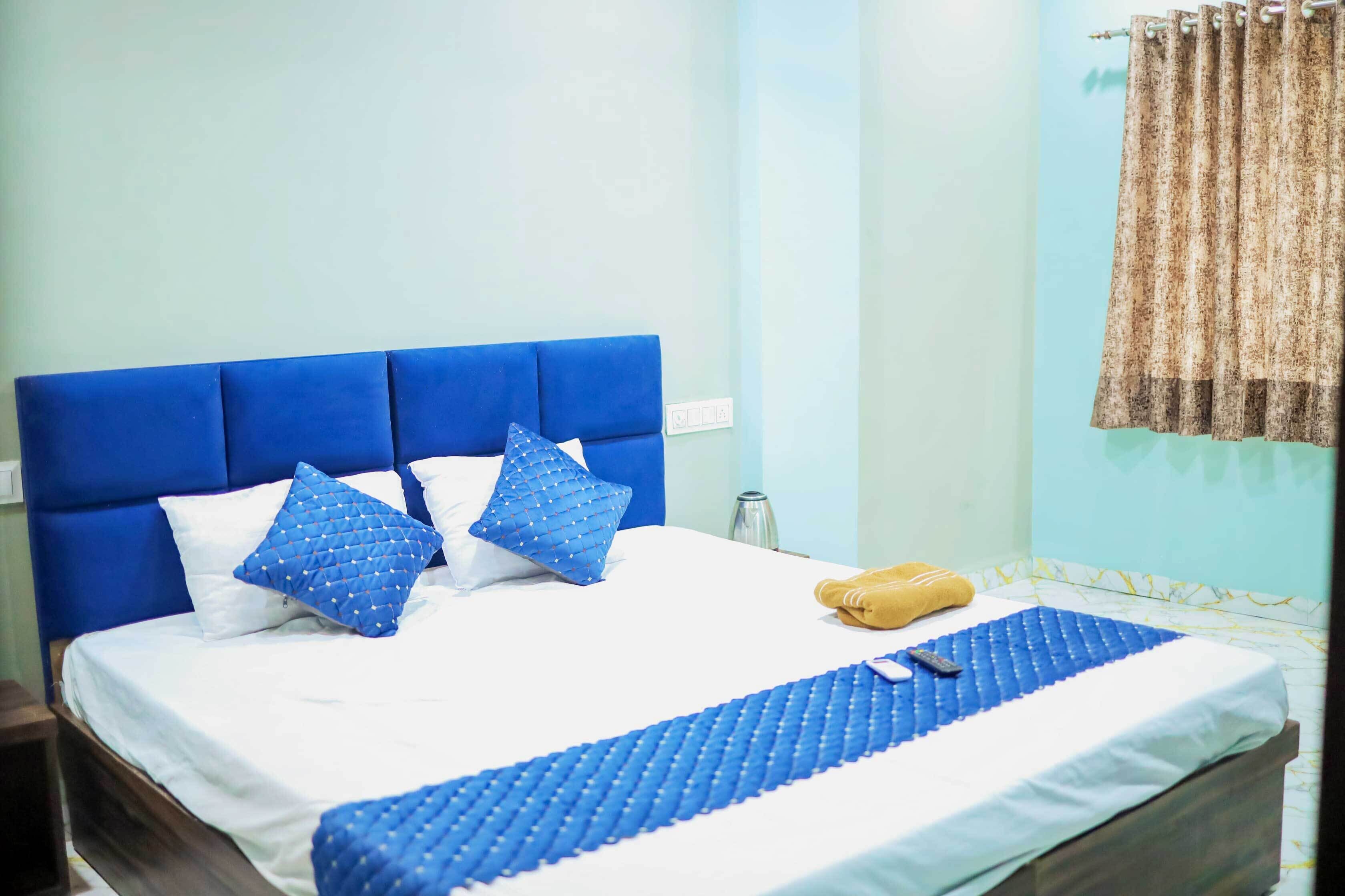 Una cama bien hecha con ropa de cama y almohadas azules, una mesita de noche con un vaso y una cortina en la ventana.