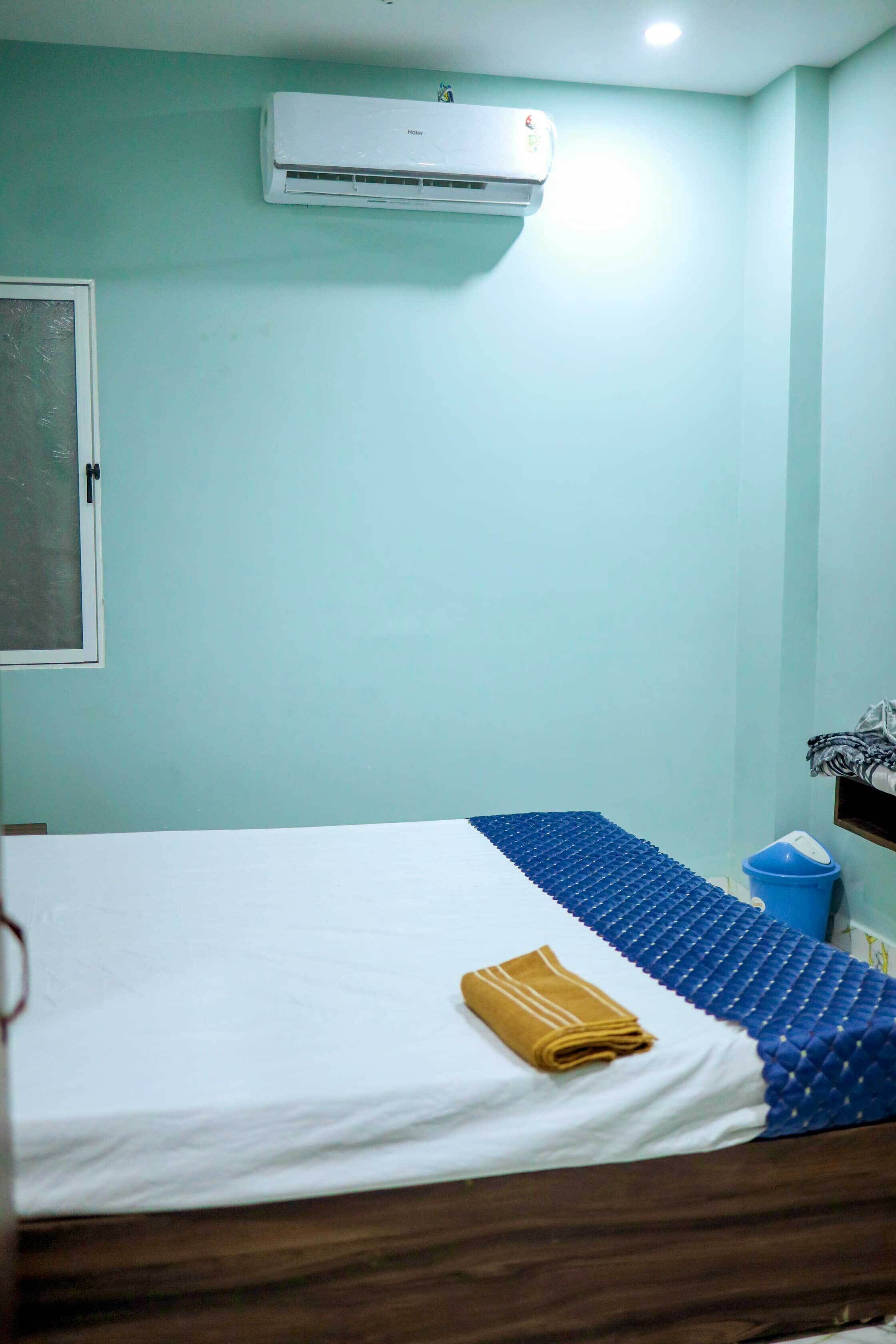 A room with a bed, a blue and white bedspread, a brown towel, an air conditioner, a window, and a blue trash bin.