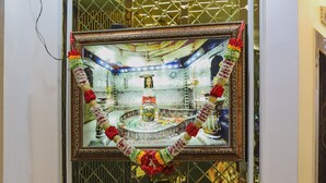 A room with a blue sofa, a framed picture on the wall, and a window with decorative elements. - Hotel Shivganga (Ujjain)