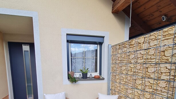 A small outdoor seating area with wicker chairs and a woven basket, a window with a view, and a stone wall. - Vacation apartment Haus-Buxbaum near Lake Wörthersee (Sankt Martin am Techelsberg)