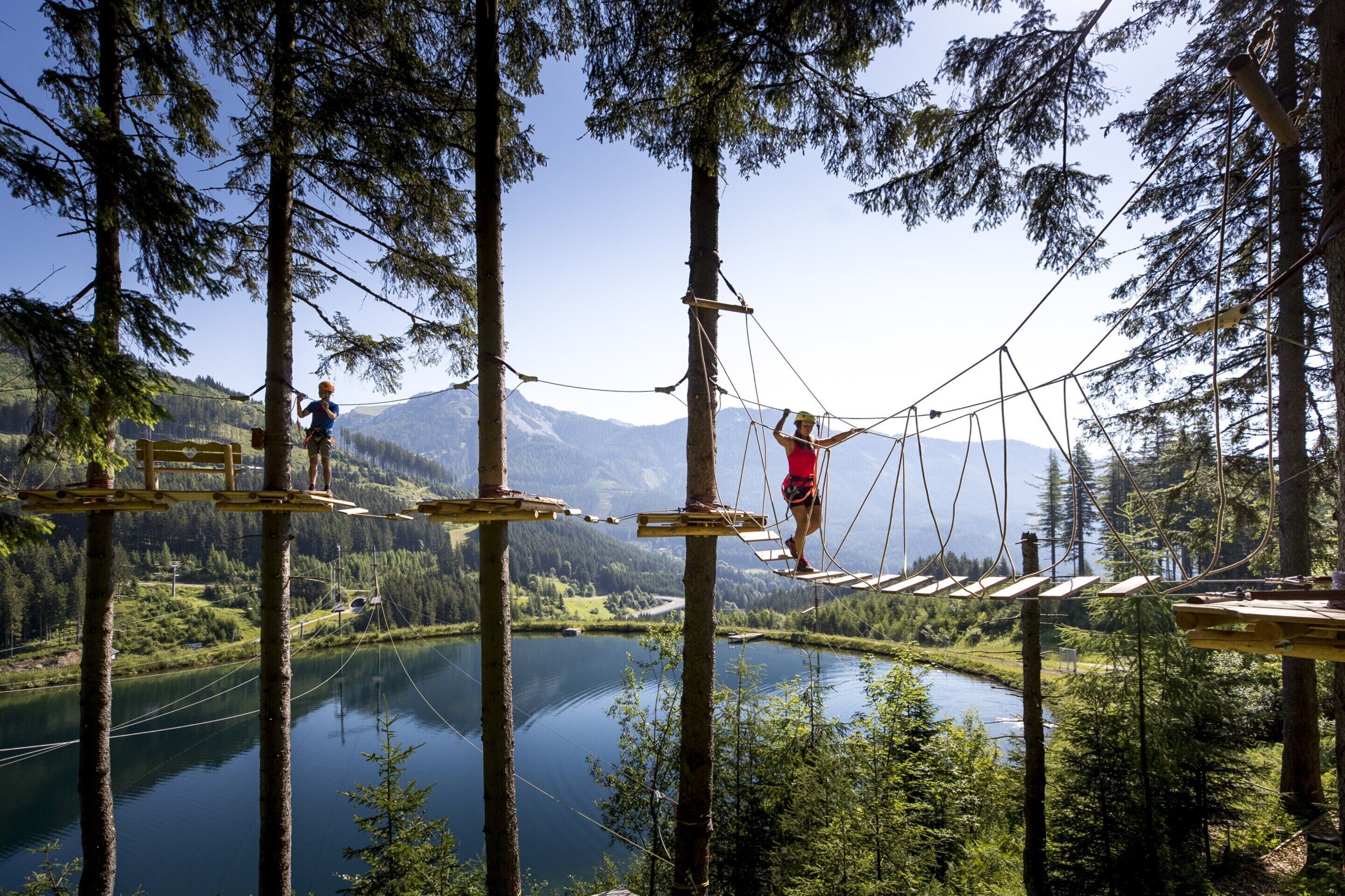 A rope bridge over a lake with forested mountains in the background.