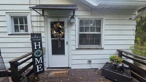Uma casa com um letreiro de boas-vindas, uma porta adornada com uma guirlanda e um pequeno alpendre com um banco e plantas em vasos.