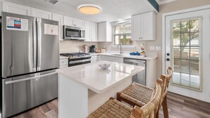 A modern kitchen with white cabinets, stainless steel appliances, a central island, and wicker chairs.