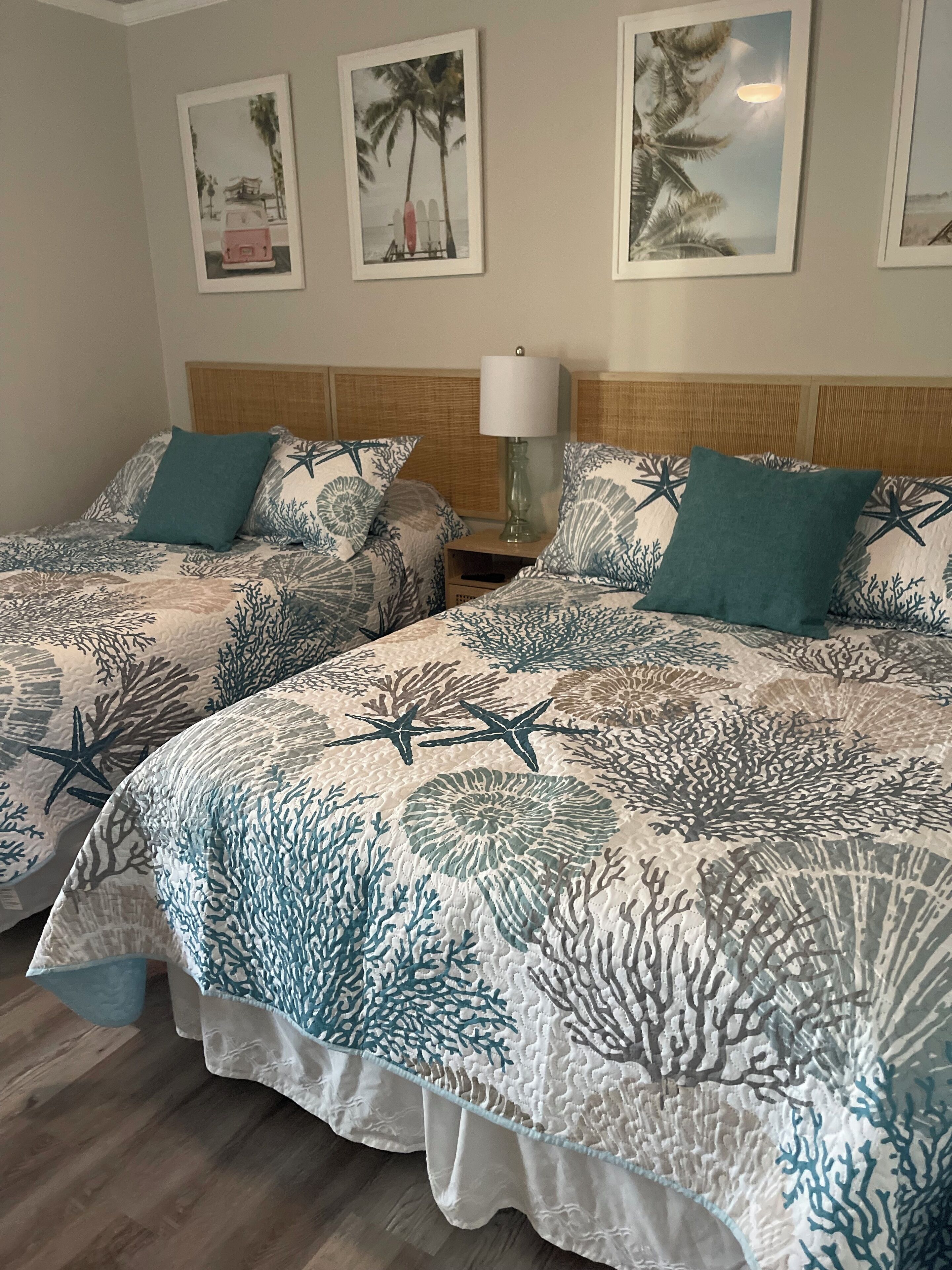 Two twin beds with coral-patterned bedding, a wooden headboard, a bedside table with a lamp, and framed pictures on the wall.