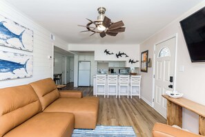 A living room with a tan sofa, a ceiling fan, and a kitchen area in the background.