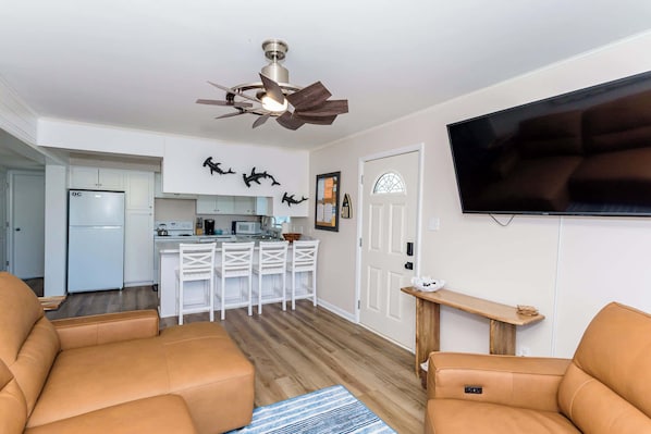 A modern living room with leather sofas, a flat-screen TV, a ceiling fan, and a kitchen in the background.
