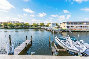 A marina with a docked speedboat, a building with a balcony, and a row of trees along the water's edge.