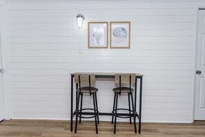 Ein Zimmer mit weißer Wandverkleidung, einem Holztisch und zwei Barhocker sowie zwei gerahmten Bildern über dem Tisch.