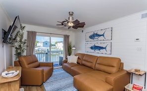 A modern living room with two brown leather sofas, a ceiling fan, and a view of a dock and houses through a sliding glass door.