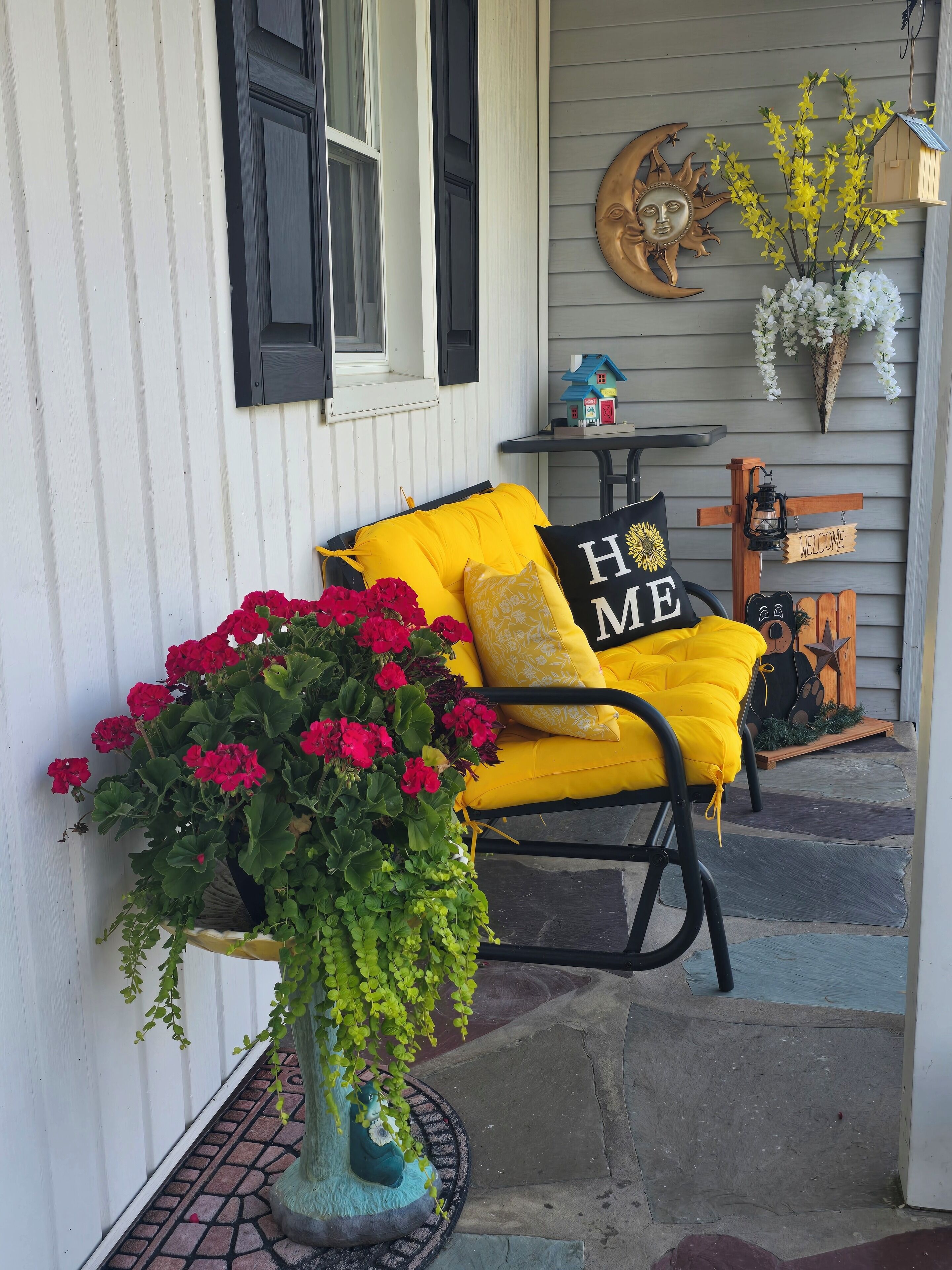 A porch with a yellow chair, a potted plant with red flowers, and a welcome sign.