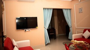 A living room with a flat-screen TV, red and white sofas, a glass coffee table, and a door leading to another room. - The Travellers Club (Guwahati)
