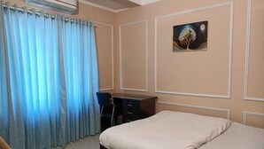 A bedroom with a bed, a desk, a chair, and an air conditioning unit. - The Travellers Club (Guwahati)