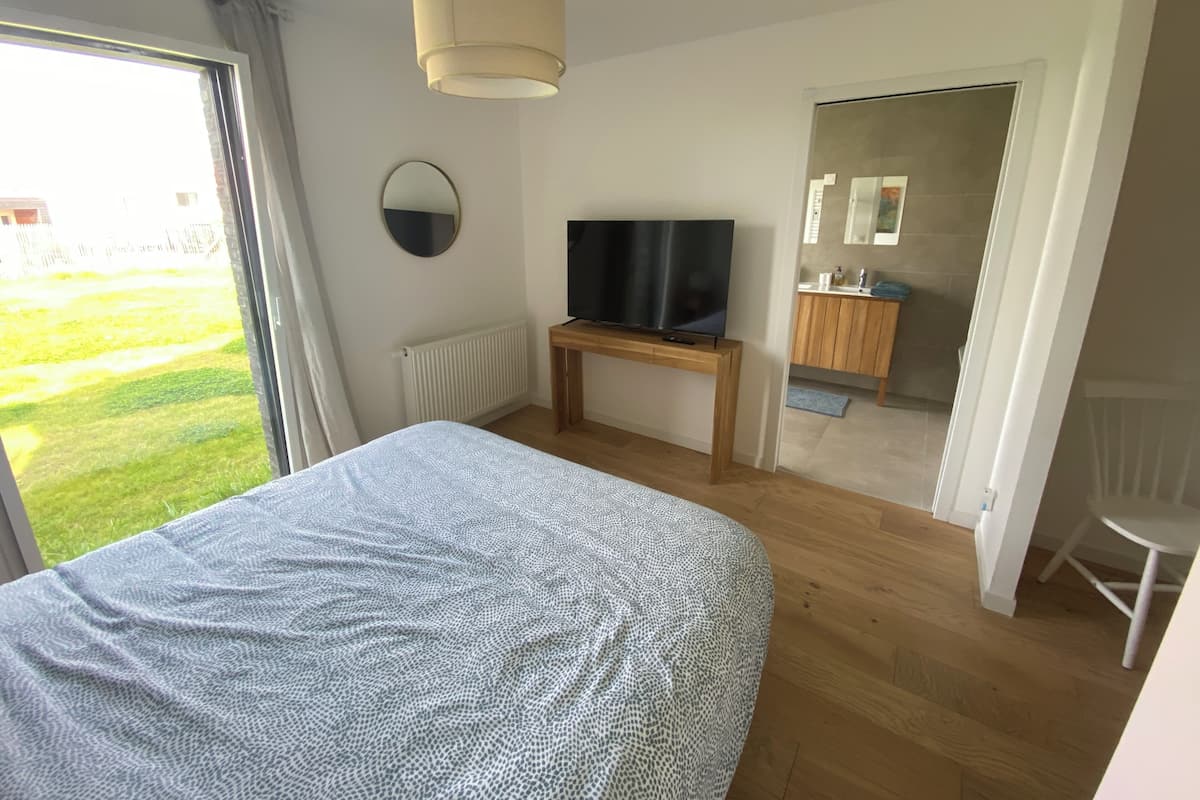Un dormitorio con cama, un televisor sobre un mueble de madera, un espejo redondo en la pared y vista a un jardín por un ventanal amplio.