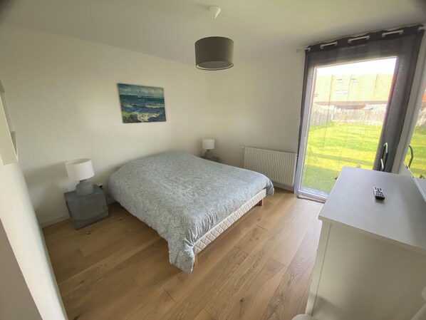A bedroom with a bed, a nightstand, a lamp, a painting, a radiator, and a view of a garden through a window.