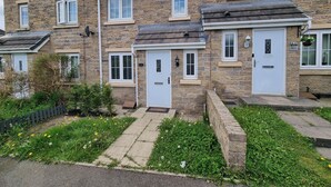 A row of stone houses with numbered doors, a paved walkway, and a garden with dandelions. - Leyland Road Retreat - 4 Bedroom House (Burnley)