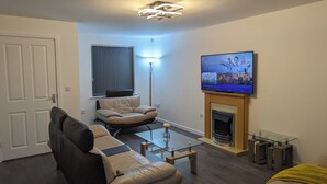 A living room with a sofa, a coffee table, a fireplace, and a TV mounted on the wall. - Leyland Road Retreat - 4 Bedroom House (Burnley)