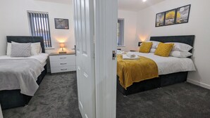 A bedroom with two beds, a nightstand, a lamp, and a window with blinds. - Leyland Road Retreat - 4 Bedroom House (Burnley)