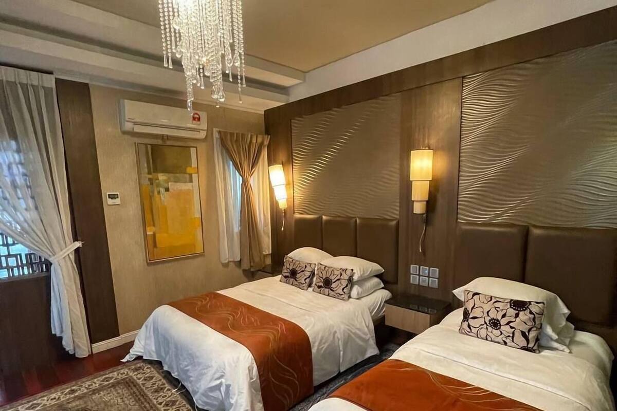 Quarto de hotel com duas camas, um candelabro, luminárias fixadas na parede, um tapete estampado e um aparelho de ar condicionado.
