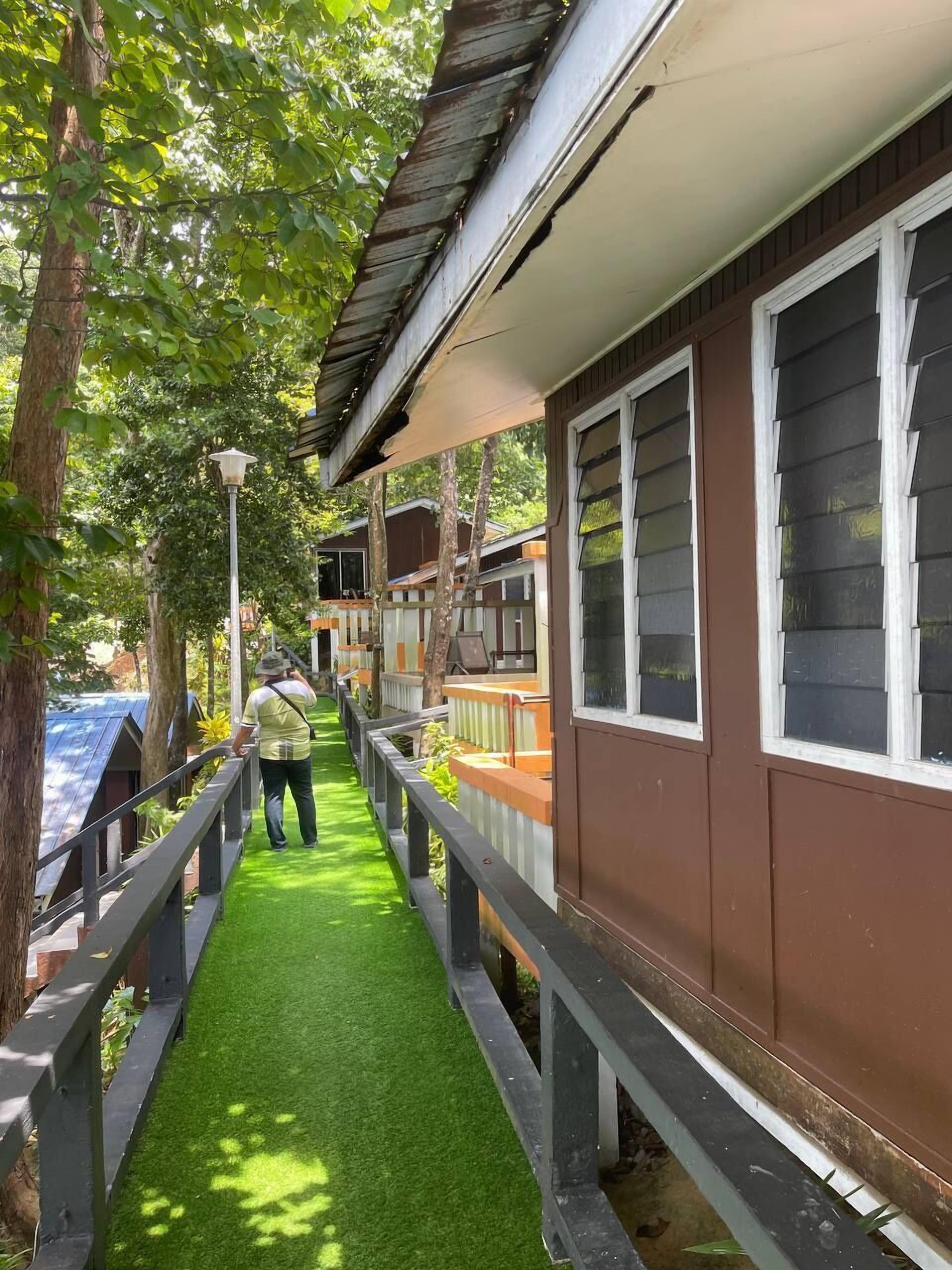 Un chemin bordé d’herbe artificielle mène à une rangée de bâtiments bruns aux encadrements de fenêtres blancs.