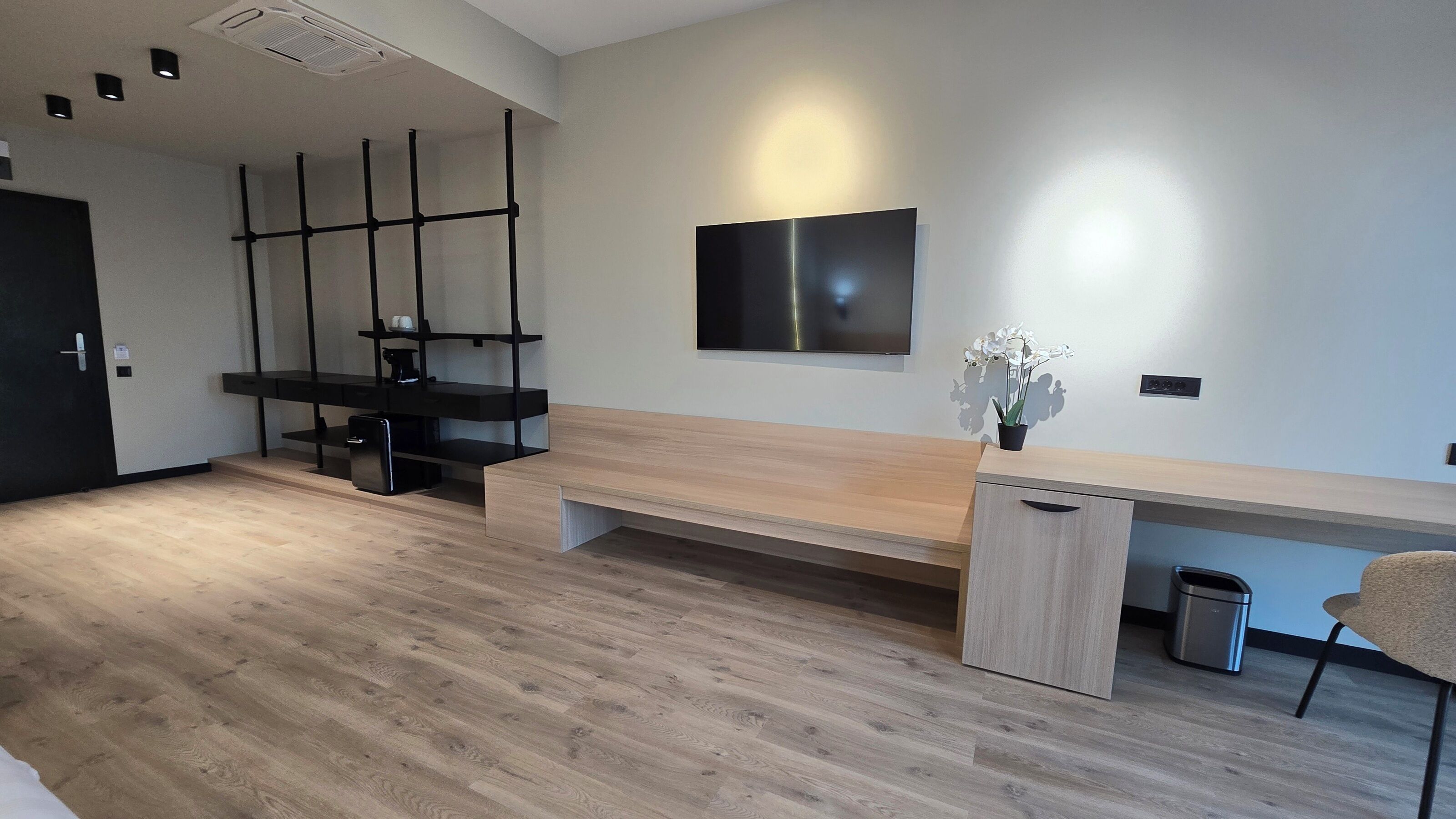 A modern, minimalist room with a wooden floor, a long desk with a built-in cabinet, a black shelving unit, and a mounted flat-screen TV.