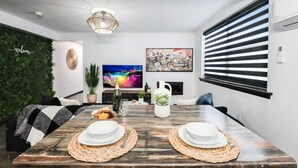 Ein stilvoller Essbereich mit einem Holztisch, gedeckt für zwei Personen, einem Fernseher und einer grünen Wand mit einem 'Welcome'-Schild.