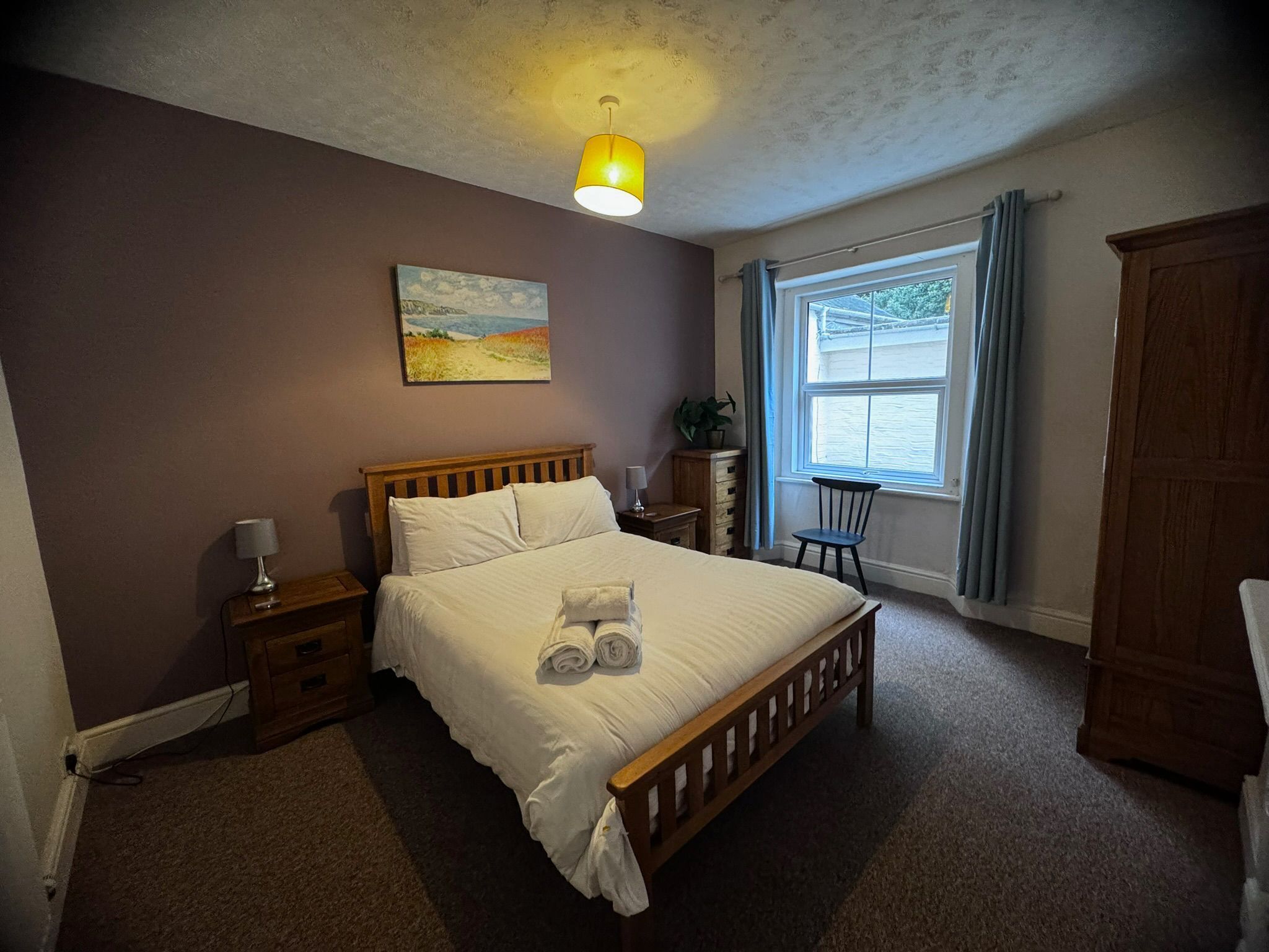Une chambre à coucher avec un lit en bois, une table de chevet, une lampe, un tableau au mur, une fenêtre avec des rideaux et une armoire.