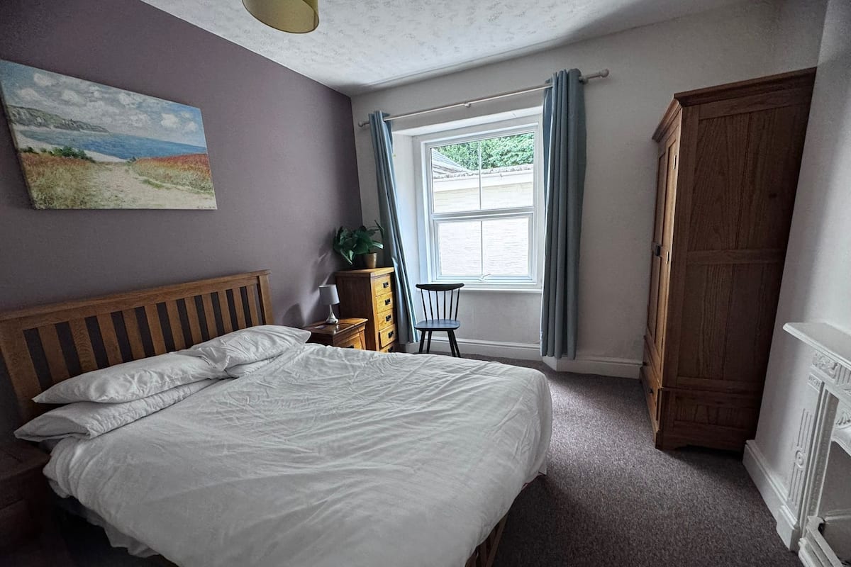 A bedroom with a wooden bed, a nightstand, a chair, a window with curtains, and a painting on the wall.