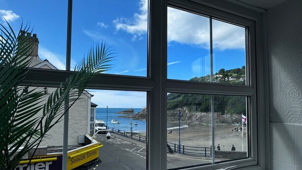 A room with large windows overlooking a coastal area, including a road with a yellow bus and a building labeled "Seaside Stores." - Seaside Apartments (Ilfracombe)
