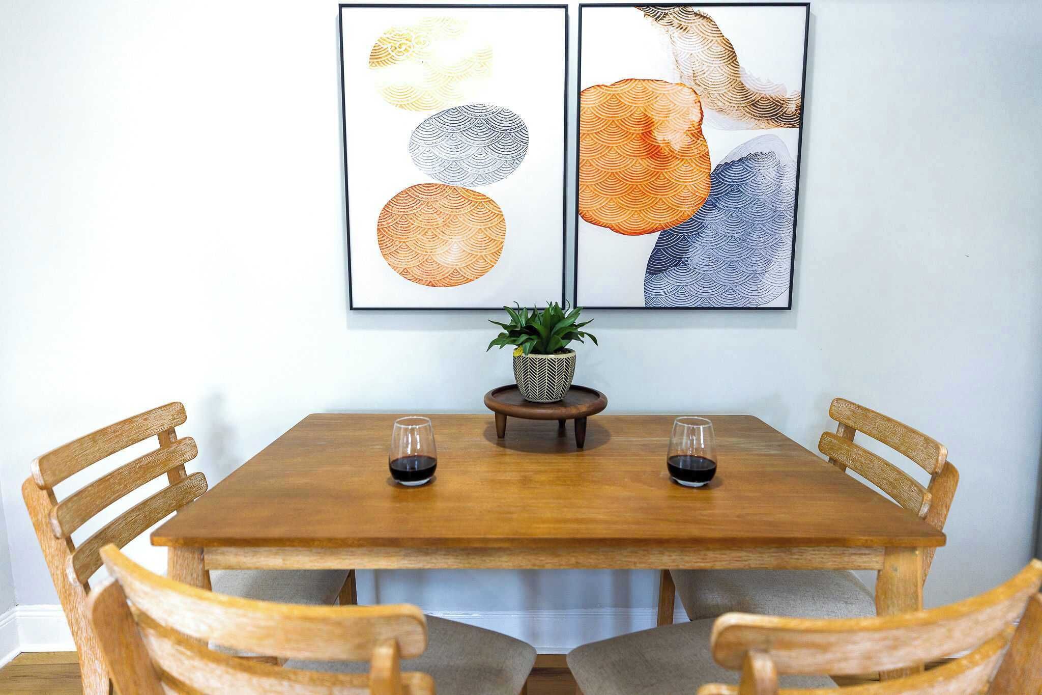 A wooden dining table with four chairs, two framed artworks above it, and a small potted plant on a wooden stand between the chairs.
