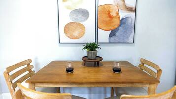 A wooden dining table with four chairs, two framed artworks above it, and a small potted plant on a wooden stand between the chairs.