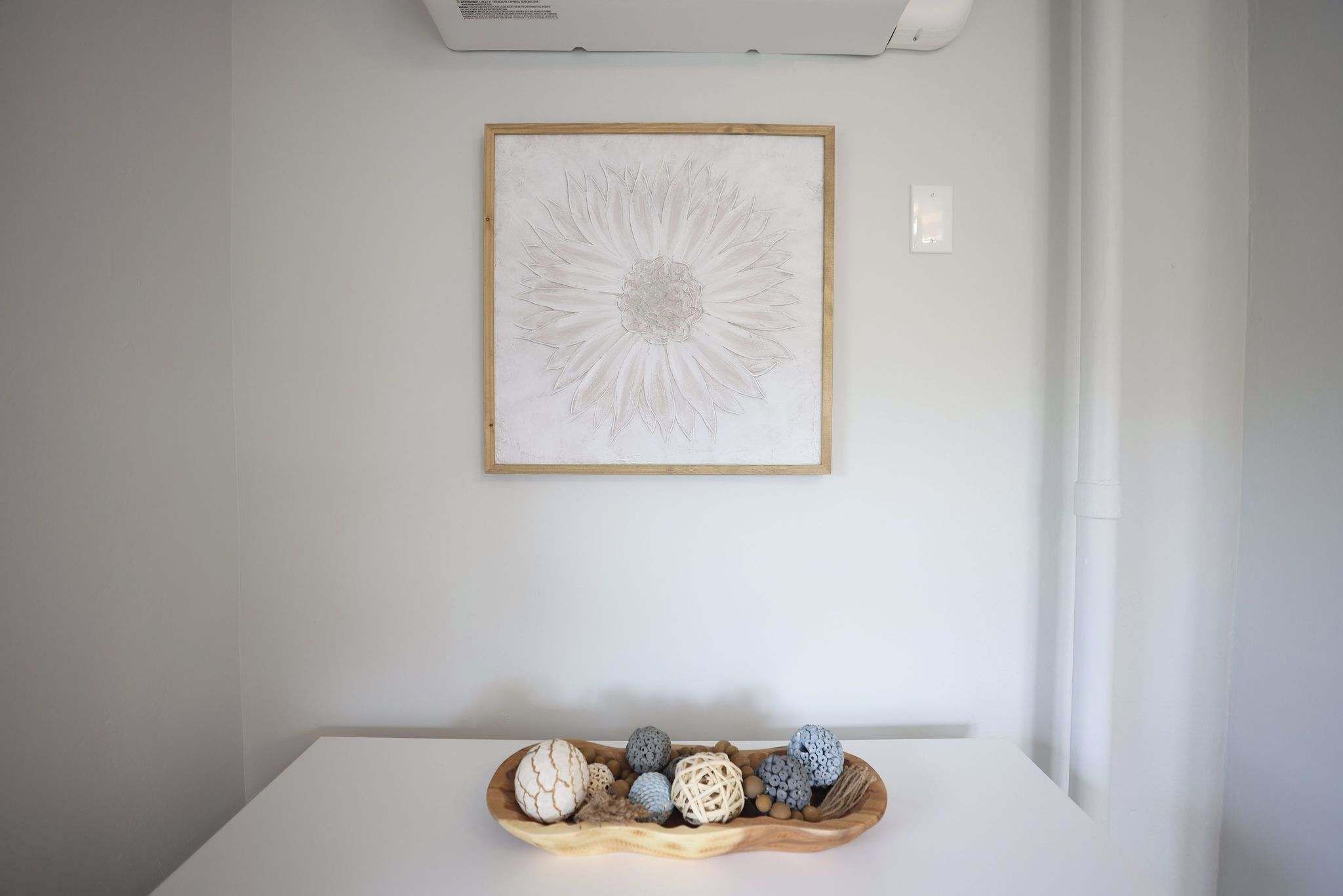 Una habitación con pared blanca, un cuadro con una flor y una bandeja de madera con objetos decorativos.