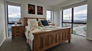Ein Schlafzimmer mit einem großen Bett, zwei Nachttischen, einem Holz-Kopfende und Blick auf eine Stadtkulisse durch große Fenster.