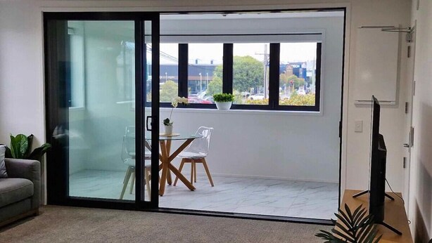 Ein modernes Wohnzimmer mit einer Schiebeglastür, die auf einen Balkon führt. Es gibt ein Sofa, einen kleinen Tisch mit Stühlen sowie ein TV-Möbel.