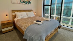 Un dormitorio moderno con una cama grande, una mesita de noche, una obra de arte abstracta en la pared y un amplio ventanal con vistas a la ciudad.
