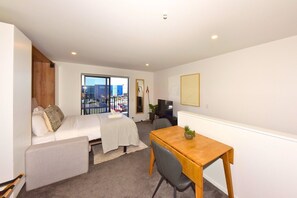 A compact room with a bed, a small table, a chair, a TV, and a window with a view of a building. - Cosy CBD Studio Apartment 12 (Christchurch)