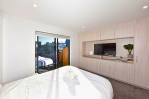 Una habitación de hotel moderna con una cama, un televisor en un mueble empotrado, una puerta corrediza de vidrio con vistas a la ciudad y una planta pequeña en el mueble.