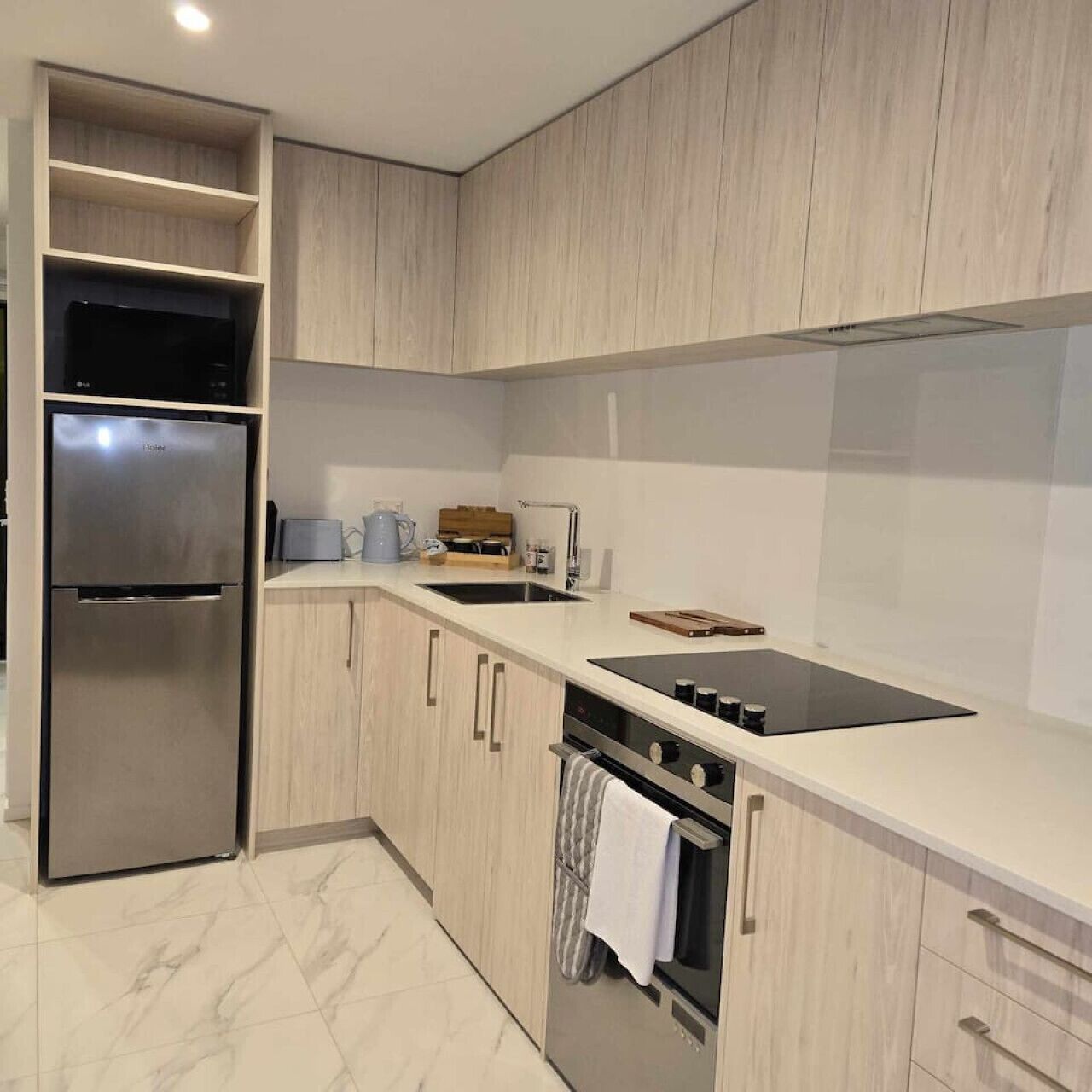 A modern kitchen with light wood cabinets, a stainless steel refrigerator, a black induction cooktop, and a built-in oven.