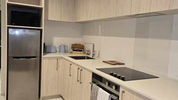 A modern kitchen with light wood cabinets, a stainless steel refrigerator, a black induction cooktop, and a built-in oven.