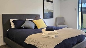 Une chambre moderne avec un grand lit, un fauteuil recouvert d’un tissu gris et une grande fenêtre.