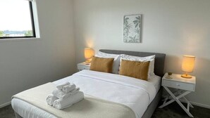 A neatly made bed with white linens and a folded towel, two bedside tables with lamps, a window with a view, and a wall-mounted artwork.