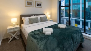 A hotel room with a large bed, two bedside tables with lamps, a view of the outside through large windows, and two framed pictures on the wall. - 10 Salisbury Cosy 2-Bed Retreat (Christchurch)