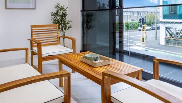 A modern interior with a wooden table and chairs, a potted plant, and a view of the street through large windows. - 10 Salisbury Cosy 2-Bed Retreat (Christchurch)