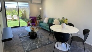 En moderne stue med en sofa, et rundt bord og et sofabord. Der er en skydedør af glas, der fører ud til en balkon med havemøbler.