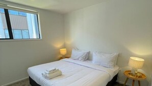 A neatly made bed with white linens and a folded towel, a bedside table with a lamp, and a window with blinds.