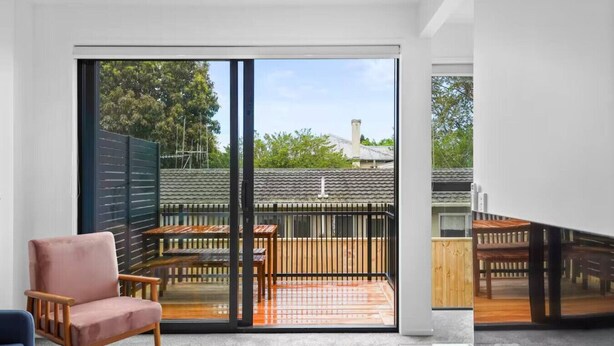 A modern living room with a sliding glass door leading to a balcony with a wooden deck and outdoor furniture. - Stunning Bay of Plenty 2 Bed (Tauranga)