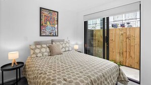A bedroom with a patterned bedspread, a nightstand with a lamp, a framed artwork on the wall, and a sliding glass door leading to an outdoor area. - Stunning Bay of Plenty 2 Bed (Tauranga)