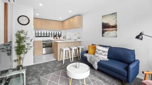 A modern living room with a blue sofa, a coffee table, and a kitchen area with wooden cabinets and a white countertop. - Stunning Bay of Plenty 2 Bed (Tauranga)