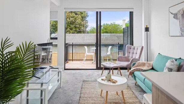 A modern living room with a sofa, armchair, and coffee table. There is a sliding glass door leading to a balcony with a view of trees and a fence. - Stylish Tauranga Home (Tauranga)