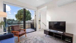 Un salon moderne doté d’un grand téléviseur, d’un canapé, d’un fauteuil et d’un balcon offrant une vue sur les arbres et les immeubles.