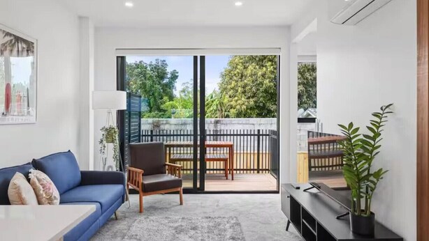 Un salon moderne avec un canapé bleu, une chaise en bois, une petite table et un meuble pour la télévision. Une porte-fenêtre coulissante donne sur une terrasse extérieure avec une plateforme en bois et une clôture en métal noir.